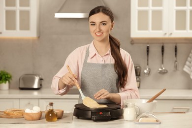 Photo of Happy woman cooking delicious crepe on electric maker at white marble table in kitchen