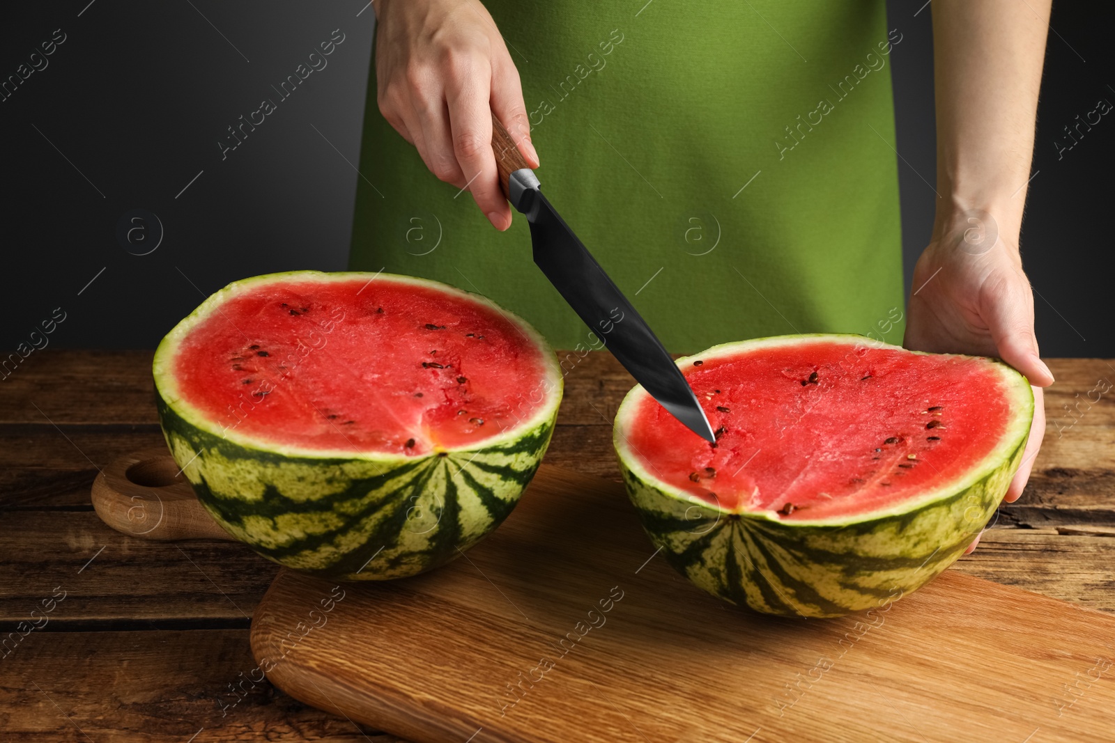 Photo of Woman cutting delicious watermelon at wooden table against dark grey background, closeup