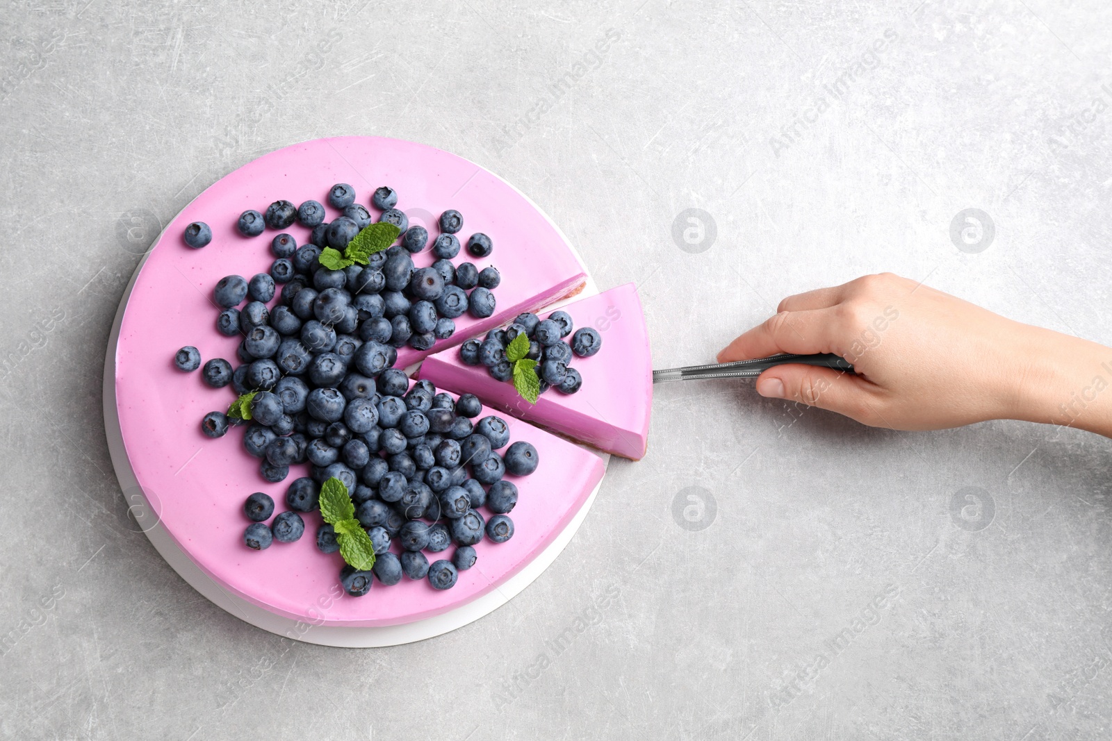 Photo of Woman taking piece of tasty blueberry cake with shovel on grey table, top view