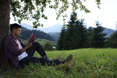 Photo of Handsome man reading book under tree on meadow in mountains