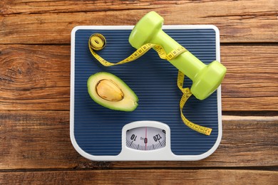 Photo of Healthy diet. Scale, avocado, measuring tape and dumbbell on wooden table, top view