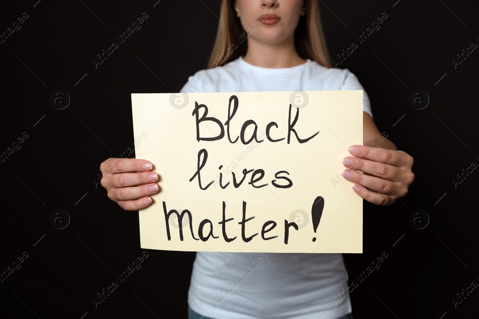 Photo of Young woman holding sign with phrase Black Lives Matter on dark background, closeup. Racism concept