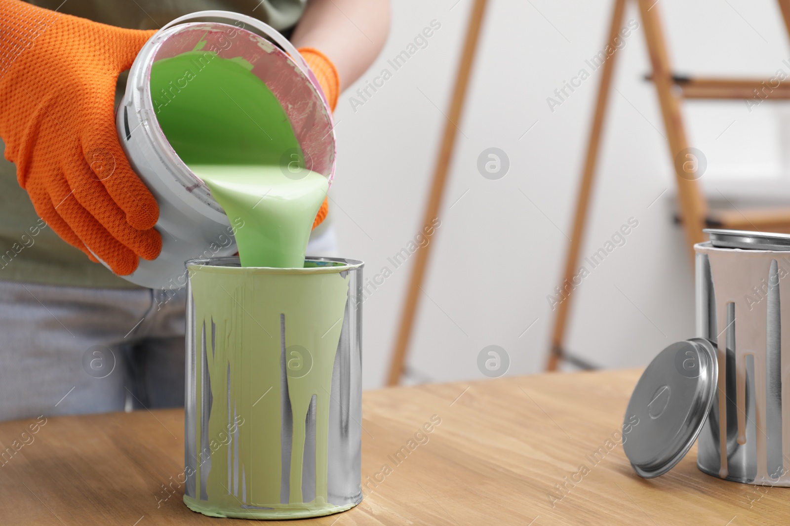 Photo of Woman pouring green paint from bucket into can at wooden table indoors, closeup