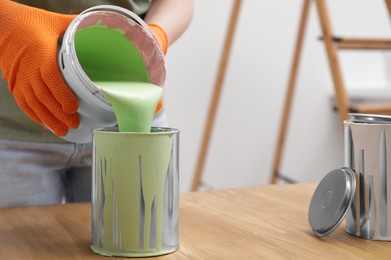 Woman pouring green paint from bucket into can at wooden table indoors, closeup