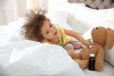 Photo of Cute African American child imagining herself as doctor while playing with stethoscope and toy bunny at home