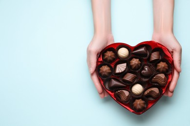 Woman holding heart shaped box with delicious chocolate candies on light blue background, top view. Space for text