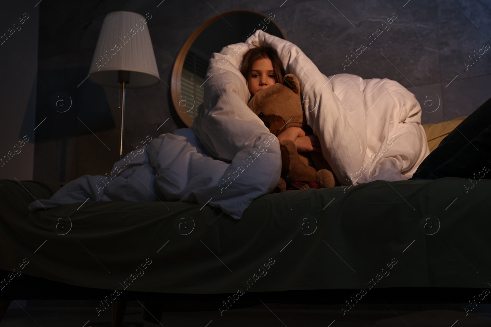 Photo of Scared little girl with toy hiding from monster in duvet at night, low angle view