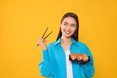 Photo of Happy young woman with plate of sushi rolls and chopsticks on orange background. Space for text
