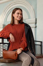 Fashionable young woman with stylish bag on bench outdoors