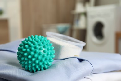 Turquoise dryer ball and detergent on clean clothes in laundry room, closeup
