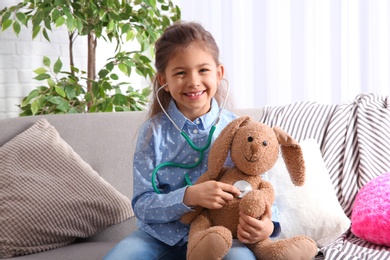 Photo of Cute child imagining herself doctor while playing with stethoscope on sofa in living room