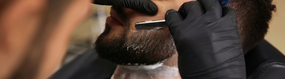 Image of Professional hairdresser shaving client with straight razor in barbershop, closeup. Banner design