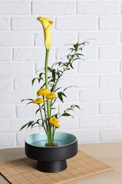 Photo of Beautiful ikebana for stylish house decor. Floral composition with fresh calla, chrysanthemum flowers and branches on wooden table near white brick wall