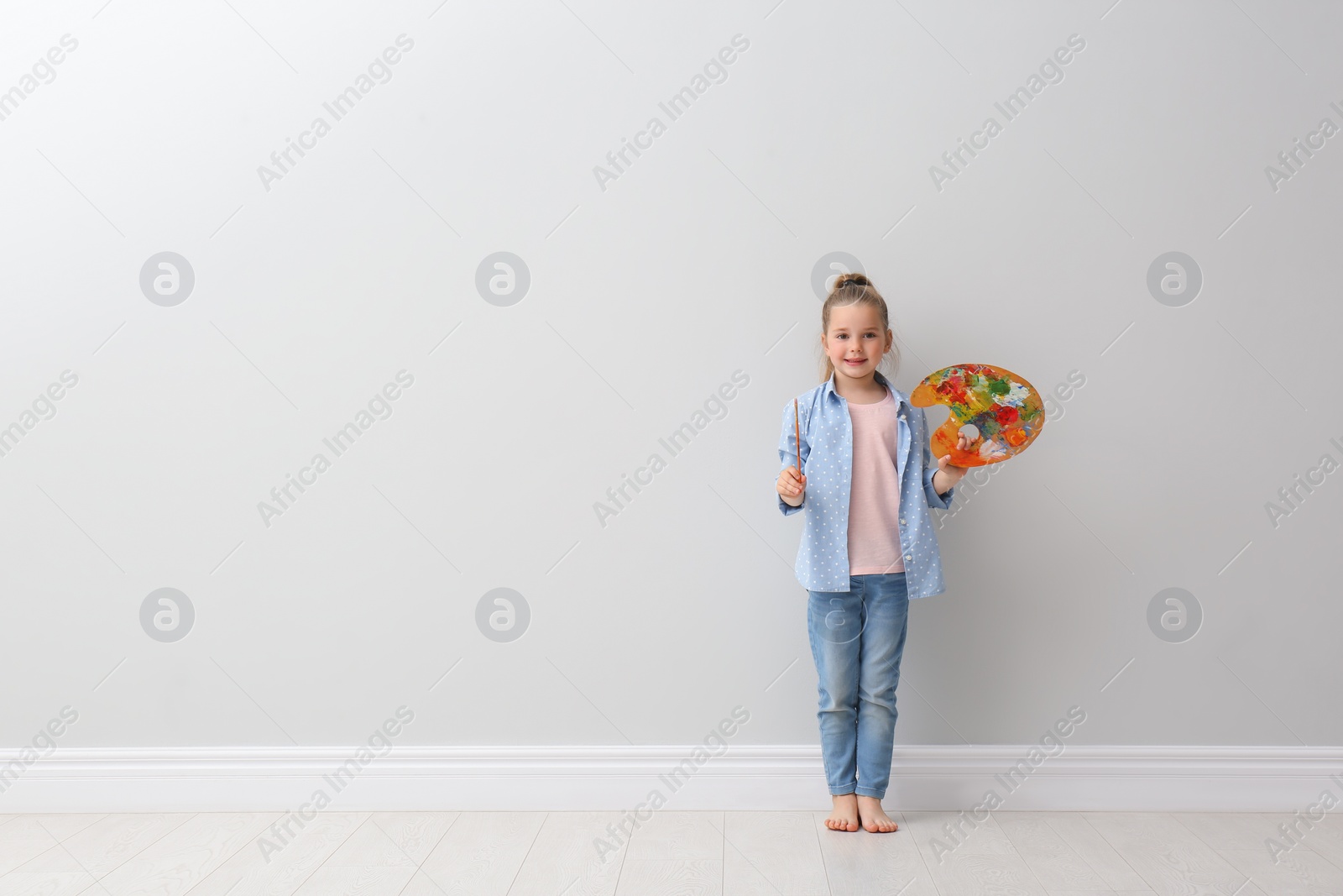 Photo of Little girl with brush and paint palette near light grey wall indoors. Space for text