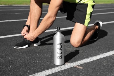 Image of Bottle with amino acids (BCAA) drink and young man tying shoelaces at stadium 