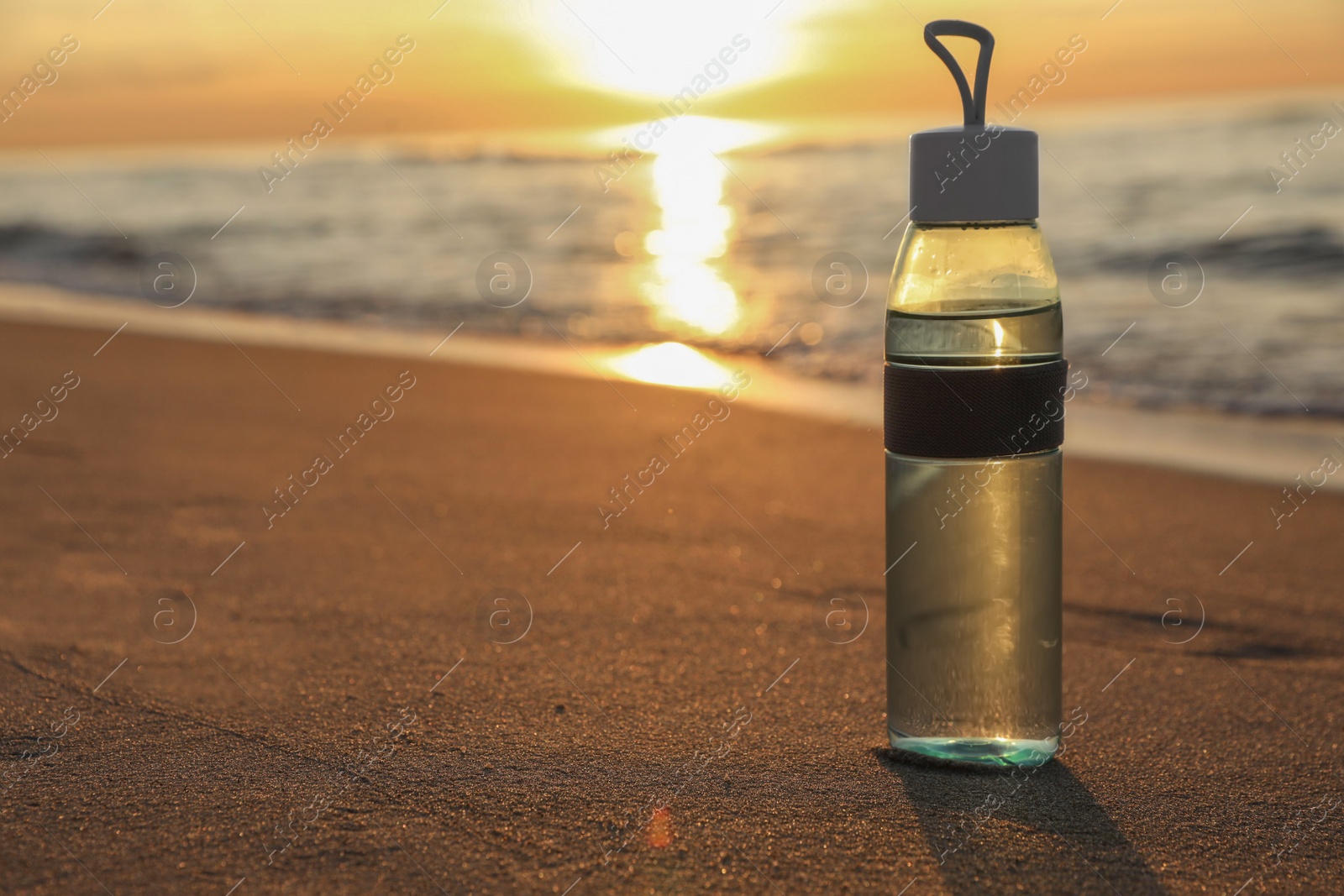 Photo of Glass bottle with water on wet sand near sea at sunset. Space for text