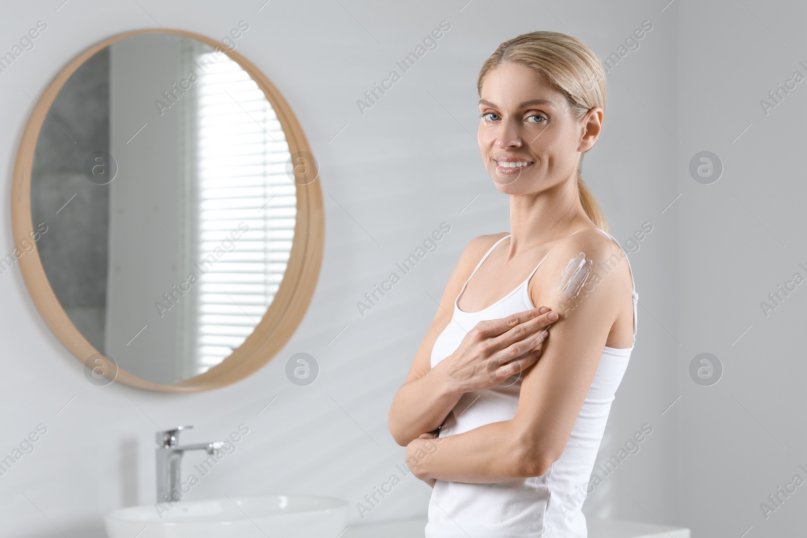 Photo of Happy woman applying body cream onto arm in bathroom, space for text
