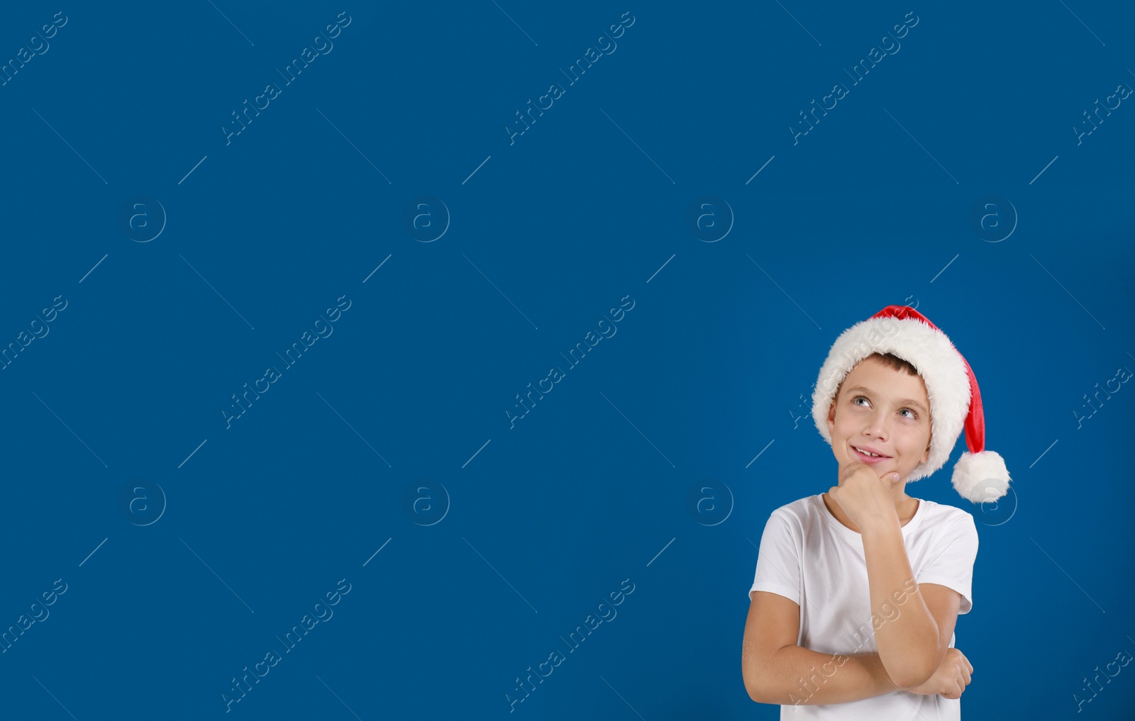 Image of Thoughtful little child in Santa hat on blue background, space for text. Christmas celebration
