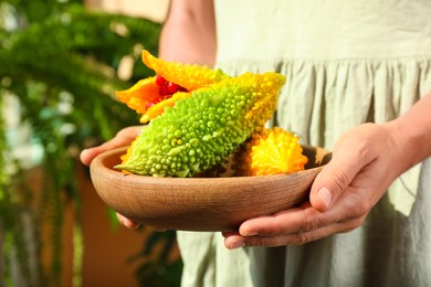 Photo of Woman holding wooden bowl with fresh bitter melons on blurred background, closeup