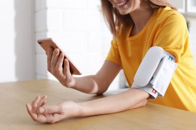 Photo of Woman checking blood pressure with modern monitor and smartphone at table indoors, closeup. Cardiology concept