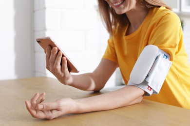 Woman checking blood pressure with modern monitor and smartphone at table indoors, closeup. Cardiology concept