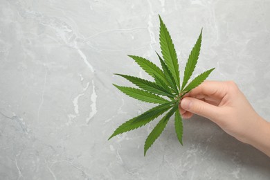 Photo of Woman holding fresh green hemp leaf at marble table, closeup. Space for text