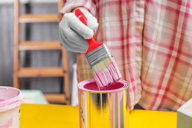 Photo of Woman dipping brush into can of pink paint at yellow table indoors, closeup