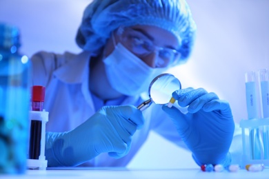 Scientist with magnifying glass examining pill at table in laboratory, closeup