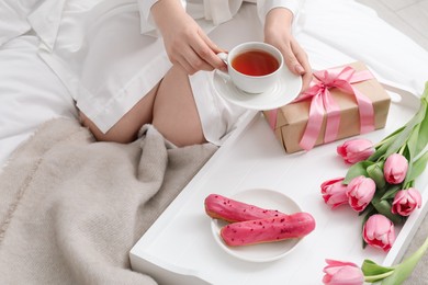 Photo of Tasty breakfast served in bed. Woman with tea, eclairs, gift box and flowers at home, closeup