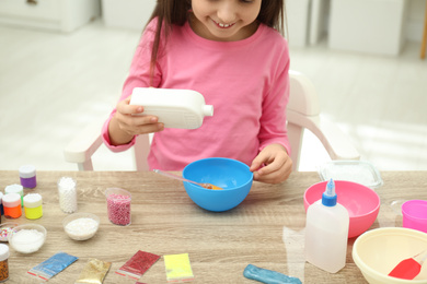 Little girl pouring glue into bowl at table in room, closeup. DIY slime toy