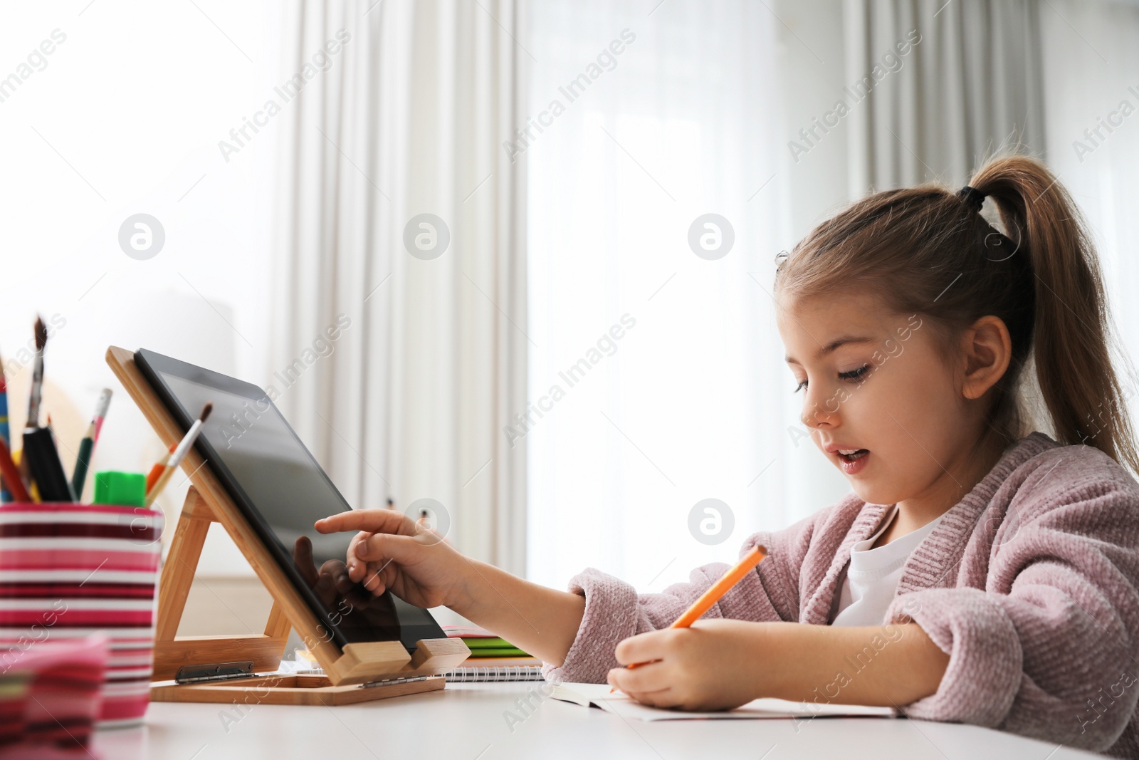 Photo of Adorable little girl doing homework with tablet at table indoors