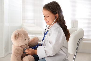 Photo of Cute little girl playing doctor with teddy bear in clinic