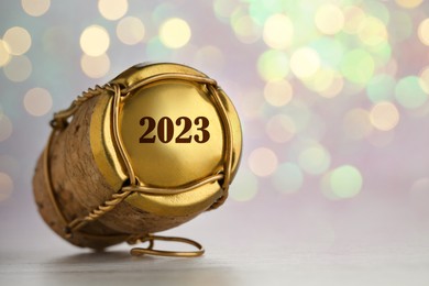 Cork of sparkling wine and muselet cap with engraving 2023 on table against blurred festive lights, closeup. Bokeh effect