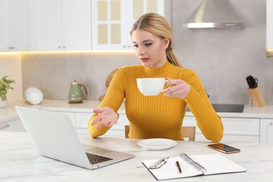 Photo of Home workplace. Woman with cup of hot drink talking by videochat on laptop at marble desk in kitchen