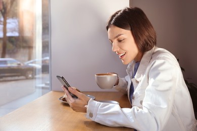 Special Promotion. Emotional young woman with cup of drink using smartphone at table in cafe