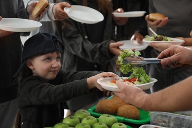 Little girl with poor people receiving food from volunteers indoors