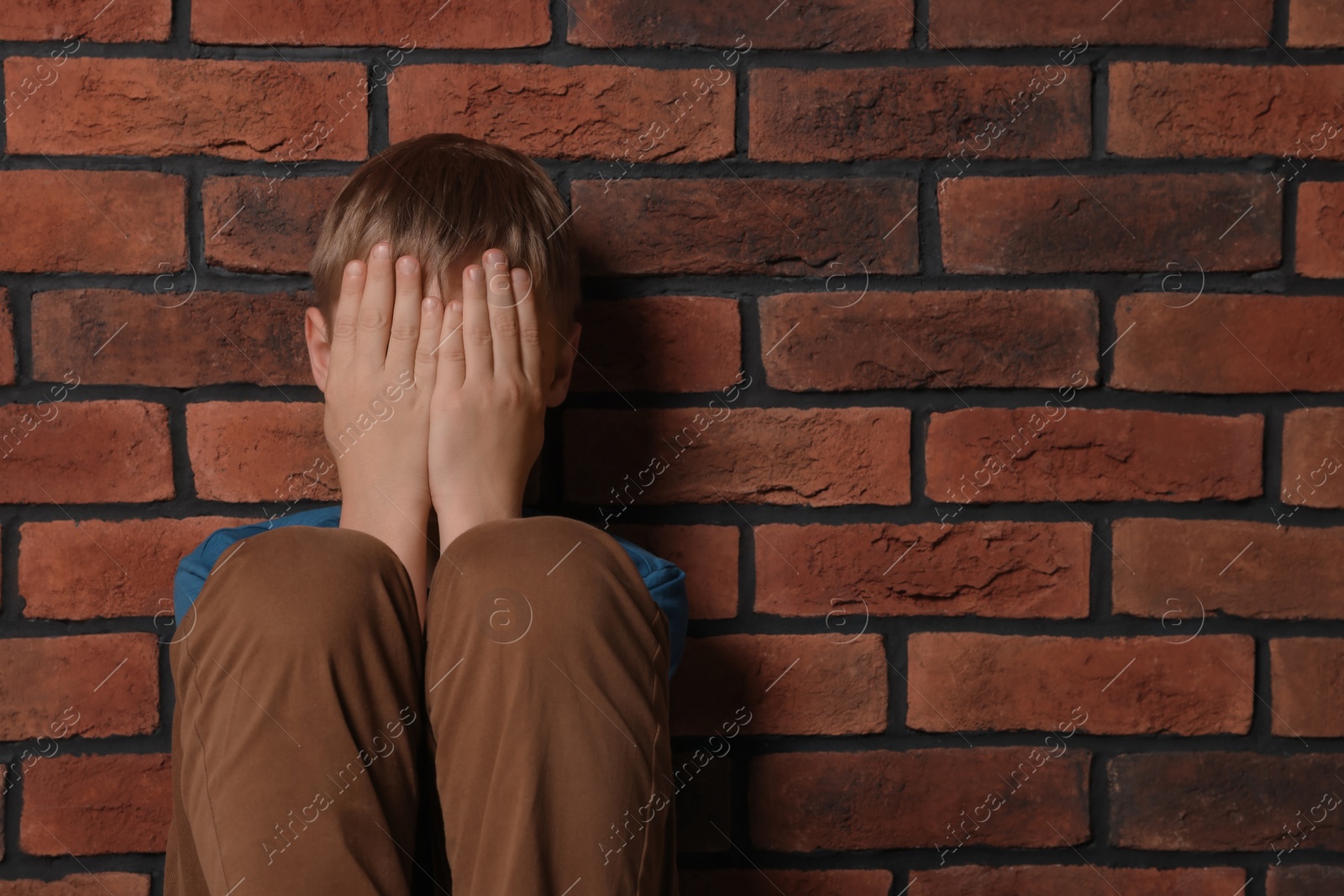 Photo of Upset boy sitting near brick wall, space for text. Children's bullying