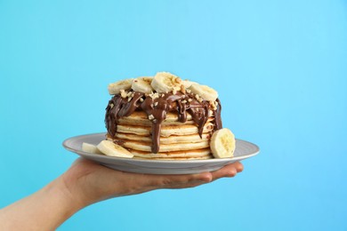Photo of Woman holding plate of tasty pancakes with chocolate spread, sliced banana and nuts on light blue background, closeup