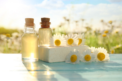 Photo of Bottles of essential oil, soap bar and fresh chamomiles on light blue wooden table in field