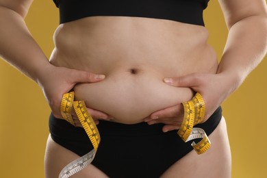 Woman with measuring tape touching belly fat on goldenrod background, closeup. Overweight problem