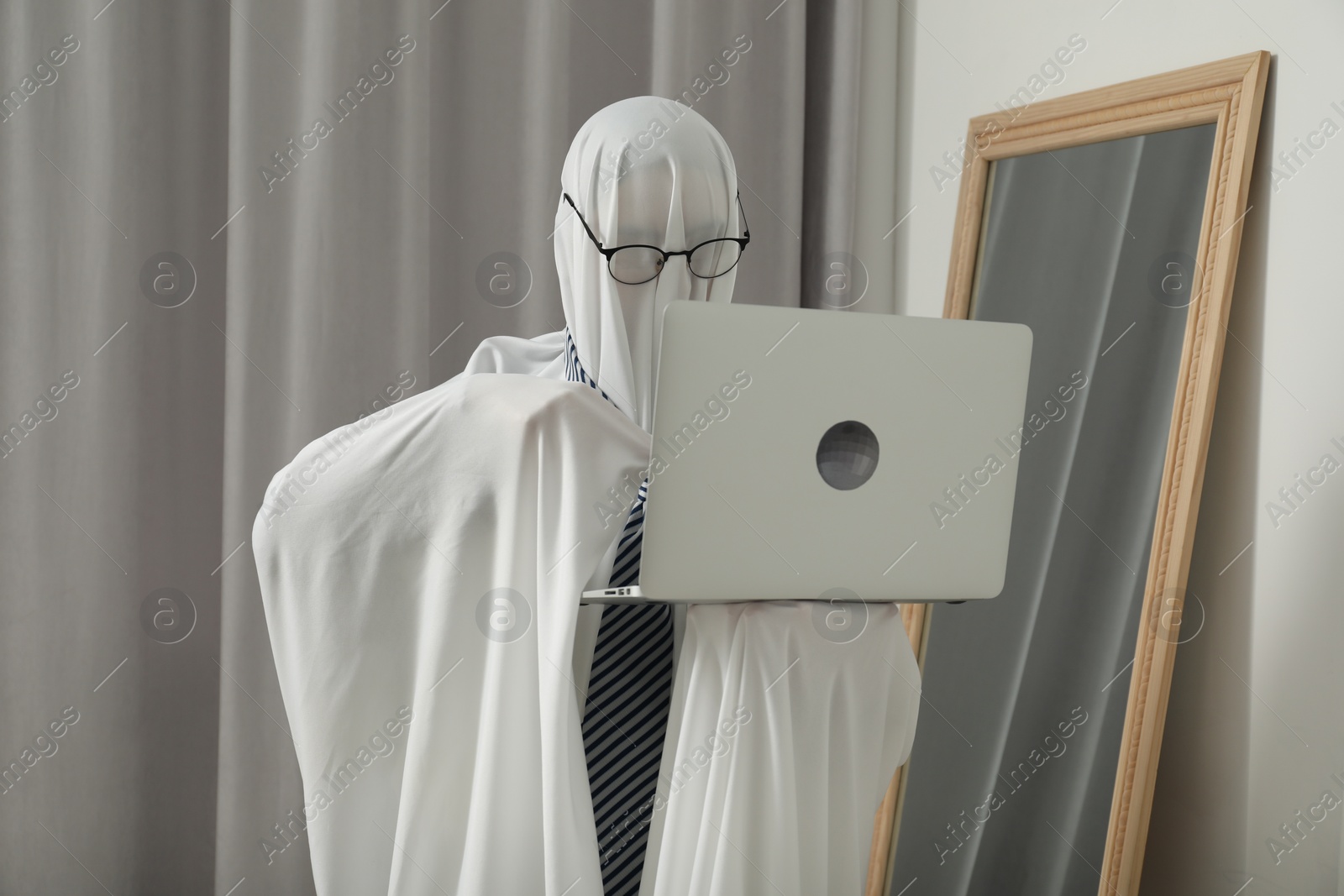 Photo of Creepy ghost. Man in white sheet using laptop at home