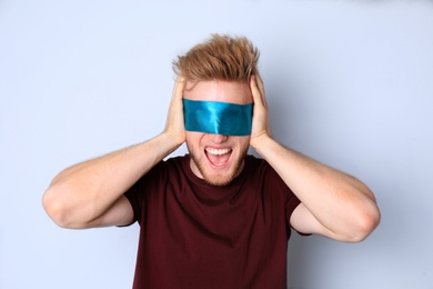 Photo of Emotional young man wearing light blue blindfold on grey background