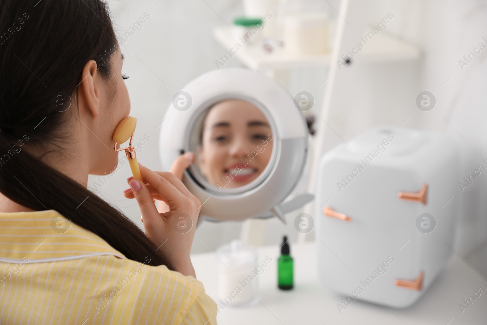 Photo of Woman doing face massage at dressing table with cosmetic refrigerator indoors