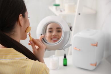 Woman doing face massage at dressing table with cosmetic refrigerator indoors