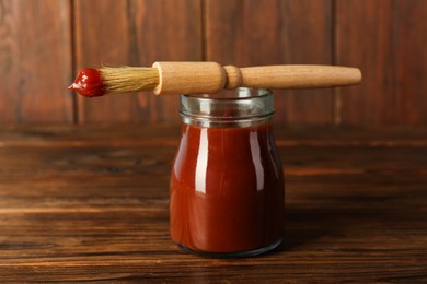 Photo of Tasty barbecue sauce in glass jar and brush on wooden table, closeup