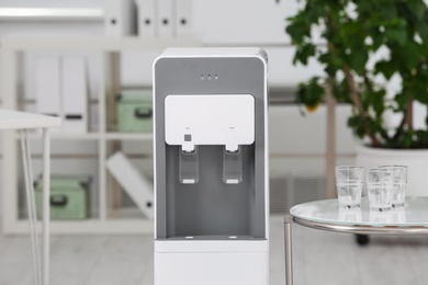 Photo of Modern water cooler and table with glasses in office