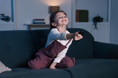Photo of Happy girl changing TV channels with remote control on sofa at home