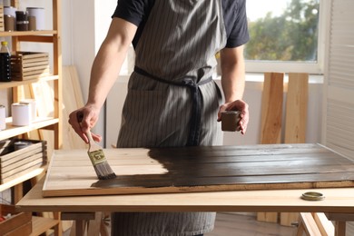 Photo of Man with brush and can applying wood stain onto wooden surface indoors, closeup