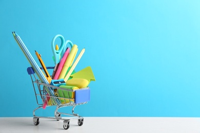 Photo of Small shopping cart with different school stationery on white table against light blue background. Space for text