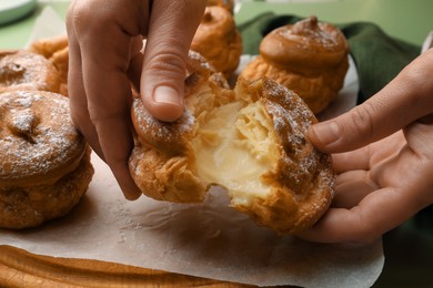 Photo of Woman breaking delicious profiterole filled with cream above wooden board, closeup